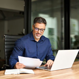 Busy middle aged professional business man using laptop computer reading document at work. Senior mature manager executive or lawyer working on laptop checking paper finance report in office. Vertical