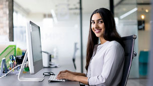 Portrait of happy young Latin-American employee wearing white shirt working at computer in office, looking at camera and smiling