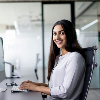 Portrait of happy young Latin-American employee wearing white shirt working at computer in office, looking at camera and smiling