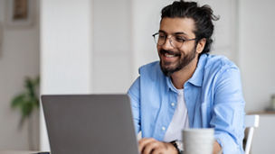 Young Indian Male Writer Using Laptop Computer, Sitting At Desk In Home Office, Handsome Western Man In Eyeglasses Typing On Keyboard And Looking At Screen, Enjoying Working Remotely, Selective Focus