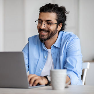 Young Indian Male Writer Using Laptop Computer, Sitting At Desk In Home Office, Handsome Western Man In Eyeglasses Typing On Keyboard And Looking At Screen, Enjoying Working Remotely, Selective Focus