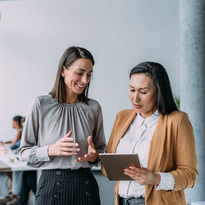 Shot of two female coworkers having a discussion in modern office. Businesswomen in meeting using digital tablet and discussing business strategy. Confident business people working together in the office. Corporate business persons discussing new project and sharing ideas in the workplace. Successful businesswomen standing with their colleagues working in background.