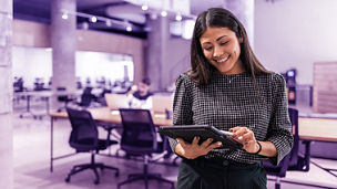 A woman in a modern office smiles as she uses a tablet, with colleagues in the background The image conveys teamwork, collaboration, and technology in a professional setting