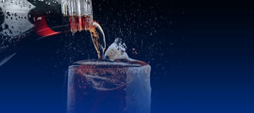 Bottle pouring a dark fizzy drink into a glass with ice, with droplets and splashes against a dark background.