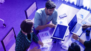 Grupo de pessoas à volta de uma mesa com diversos papéis com gráficos, portáteis e com um tablet. Um dos colegas apresenta os dados do tablet aos restantes.