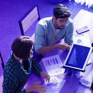 Grupo de pessoas à volta de uma mesa com diversos papéis com gráficos, portáteis e com um tablet. Um dos colegas apresenta os dados do tablet aos restantes.