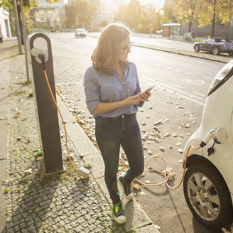 Young woman is standing near the electric car and looks at the smart phone. The rental car is charging at the charging station for electric vehicles. Car sharing.