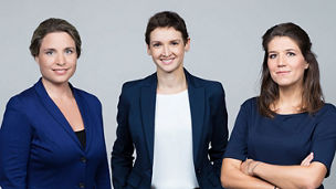 Das Bild zeigt drei Personen in formeller Kleidung, die vor einem neutralen Hintergrund stehen.