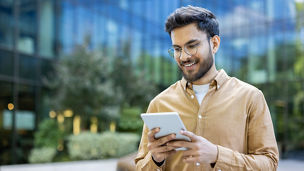 Young man smiling while using tablet outside modern office building. Casual attire, urban setting, and contemporary design. Represents technology, business, and communication. Happy expression