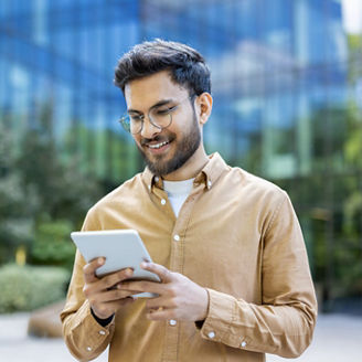 Young man smiling while using tablet outside modern office building. Casual attire, urban setting, and contemporary design. Represents technology, business, and communication. Happy expression