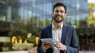 Young businessman in suit using tablet outside modern glass office building. Confident professional smiling working on digital device. Concept of business, technology, success, and entrepreneurship.