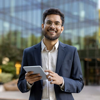 Young businessman in suit using tablet outside modern glass office building. Confident professional smiling working on digital device. Concept of business, technology, success, and entrepreneurship.