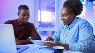 Two casually dressed African colleagues sitting together at a table in a modern office working with a digital tablet and laptop