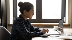 Distant working with finance. Thoughtful young mixed race female sitting by desk at home focused on pc screen studying statistic documents. Indian lady remote employee pondering on investment research