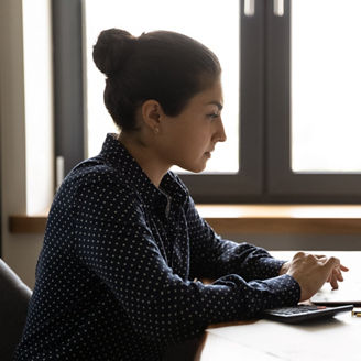 Distant working with finance. Thoughtful young mixed race female sitting by desk at home focused on pc screen studying statistic documents. Indian lady remote employee pondering on investment research