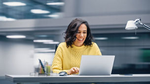 Modern Office: Happy Smiling Hispanic Businesswoman Sitting at Her Desk Working on a Laptop Computer Celebrates Victory. Latin Female Entrepreneur is a Happy Winner. Motion Blur Background