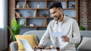 Indian young man working with documents and bills at home, sitting on sofa. holds papers and uses a laptop.