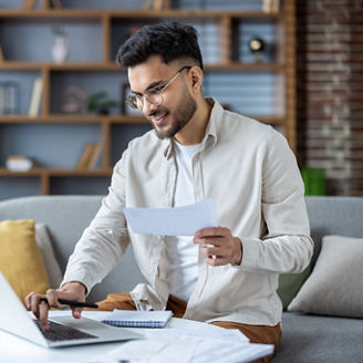 Indian young man working with documents and bills at home, sitting on sofa. holds papers and uses a laptop.