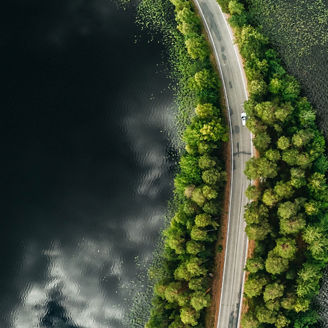 Eine Luftaufnahme einer Straße, die sich durch eine schmale Landzunge mit grüner Vegetation schlängelt, umgeben von einem dunklen Gewässer. Ein weißes Auto fährt auf der Straße.