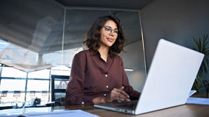 Young professional it specialist latin hispanic business lady working on laptop pc sitting at desk in modern office space. 30s middle eastern indian woman using computer technology app for work online