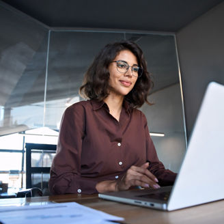 Young professional it specialist latin hispanic business lady working on laptop pc sitting at desk in modern office space. 30s middle eastern indian woman using computer technology app for work online