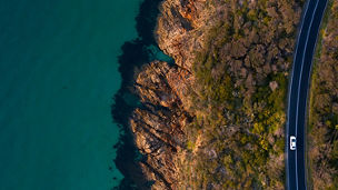 Eine Luftaufnahme einer Küstenstraße, die entlang einer felsigen Küste verläuft, mit einem Auto darauf und dem Meer auf der linken Seite.