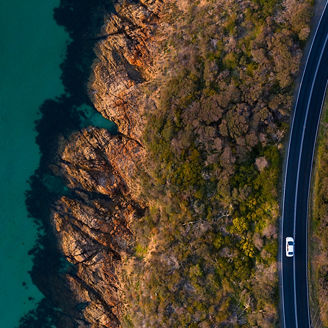 Eine Luftaufnahme einer Küstenstraße, die entlang einer felsigen Küste verläuft, mit einem Auto darauf und dem Meer auf der linken Seite.