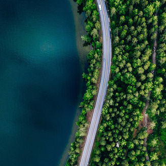 Eine Luftaufnahme einer Straße, die sich durch einen dichten Wald schlängelt und an einem großen Gewässer entlangführt.