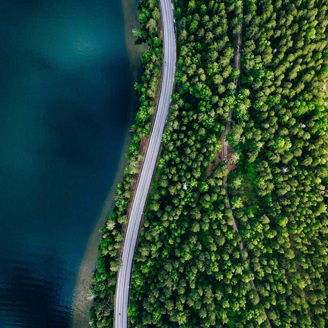 Eine Luftaufnahme einer Straße, die sich durch einen dichten Wald schlängelt und an einem großen Gewässer entlangführt. Die Straße verläuft durch eine schmale Landzunge, die von Wasser auf beiden Seiten umgeben ist.