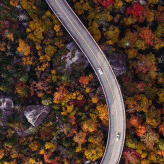 Eine Luftaufnahme einer kurvigen Straße, die sich durch einen Wald mit Herbstlaub schlängelt. Die Bäume sind in leuchtenden Farben wie Rot, Orange und Gelb gefärbt. Ein weißes Auto fährt auf der Straße, die von Felsen und bunter Vegetation umgeben ist.