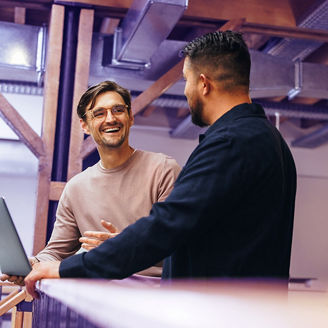 Two business men talking to each other about their tech work. Business colleagues discussing efficient ideas to finish a project. Male professionals working in a startup.; Shutterstock ID 2300432543; purchase_order: advisory assurance marketing; job: anne; client: ai-hub