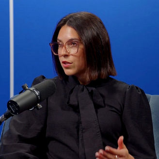 Person speaking into a microphone in a studio setting, seated against a blue background with vertical light accents
