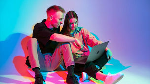 Stylish young couple with laptop working, watching series or shopping online. Colorful neon light. High quality photo