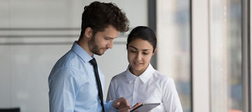 Two busy diverse young business coworkers in formalwear using tablet computer, watching online presentation. Employees discussing startup project. Indian professional consulting colleague expert