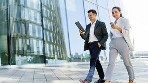 Two business professionals walking confidently outside a modern office building, engaged with a tablet and smartphone.

