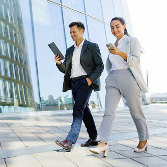 Two business professionals walking confidently outside a modern office building, engaged with a tablet and smartphone.

