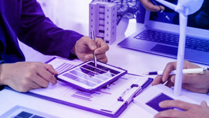 Numa mesa de escritório veem-se mãos de várias pessoas a mostrar dados em tablet e portátil enquanto que uma mão segura um protótipo de uma turbina eólica