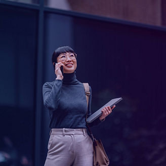 Person standing outdoors in front of a glass building, holding a tablet and talking on a mobile phone