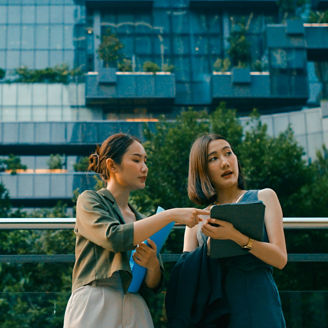 Two people standing outdoors in front of a modern glass building, looking at and discussing information on a tablet.