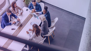 Group of business professionals seated around a conference table collaborating on laptops, documents, and charts in a modern office setting.