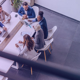 Group of business professionals seated around a conference table collaborating on laptops, documents, and charts in a modern office setting.