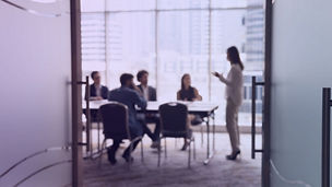 View into a conference room where a group of people sit around a table while another person stands and presents in front of large windows overlooking the city.