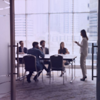 View into a conference room where a group of people sit around a table while another person stands and presents in front of large windows overlooking the city.