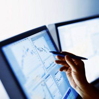 Close-up of a hand holding a pen pointing at financial charts on a computer monitor