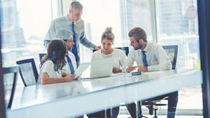 Group of business people working. They are at the boardroom table. They are all well dressed men and women. They have a laptop computer. City background