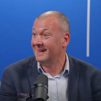 Person speaking into a microphone in a studio, seated against a blue background with vertical light accents