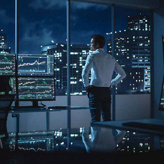 Successful Businessman Looking Out of the Window on Late Evening. Modern Hedge Fund Office with Computer with Multi-Monitor Workstation with Real-Time Stocks, Commodities and Exchange Market Charts.