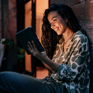Smiling woman holds tablet while sitting against wall, enjoying remote connection with warmth and ease