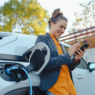 Young woman with smartphone waiting while her electric car charging in home charging station, sustainable and economic transportation concept.