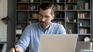 Confident businessman wearing glasses writing notes or financial report, sitting at desk with laptop, focused serious man working with paper documents, student studying online, research work
